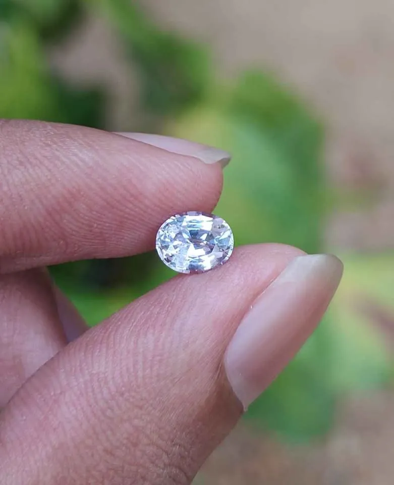 Person holding a faceted oval light pink Taaffeite gemstone between fingers with a natural background.