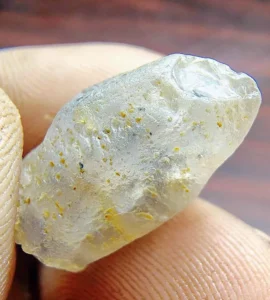 A close-up macro photograph of a rough, unpolished white sapphire crystal with minor yellowish natural inclusions held between fingertips.