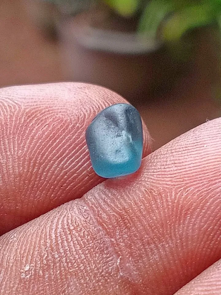 A small rough natural blue spinel gemstone held between fingertips.