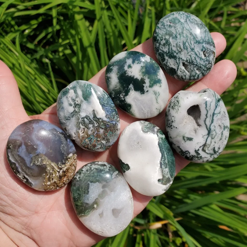 A hand holding a collection of smooth, polished moss agate palm stones displaying distinct green moss-like inclusions and natural druzy crystal cavities against lush outdoor greenery.