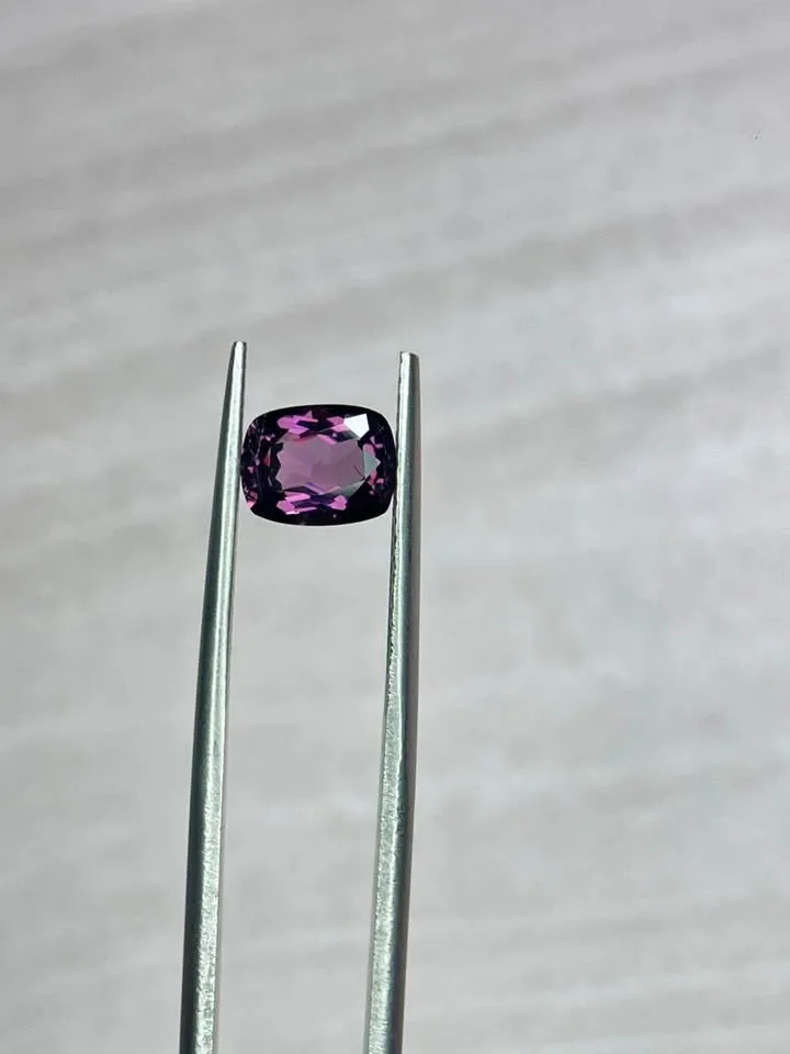 A faceted, cushion-cut purple spinel gemstone held securely with metal jewelers tweezers against a neutral background.