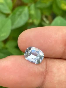 A faceted cushion-cut white sapphire resting on fingertips outdoors against a soft green leafy background.