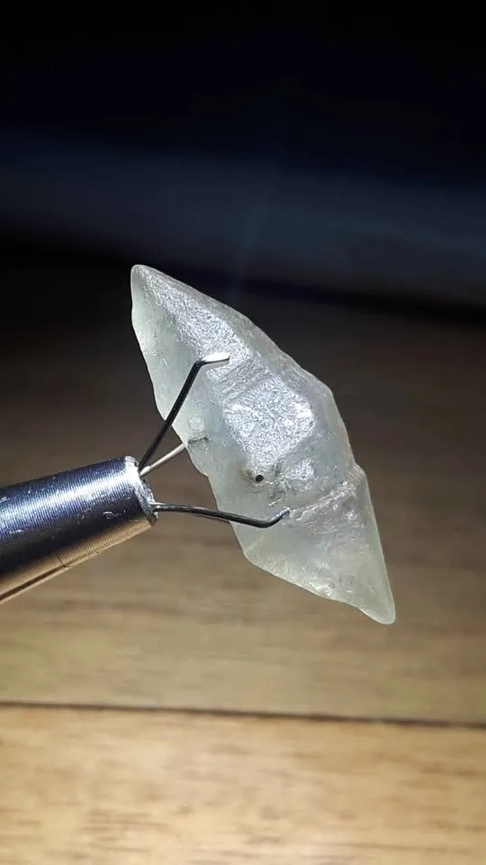 A close-up view of a raw, uncut white sapphire crystal with a pointed natural form held in a gem holder tool.