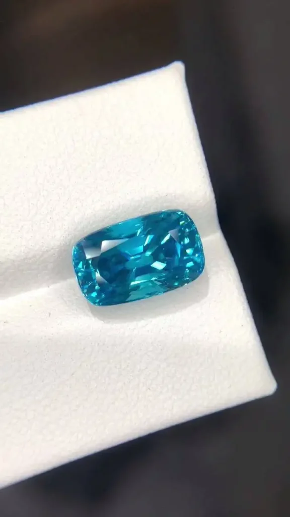 A precision-faceted cushion-cut blue zircon gemstone displaying vivid electric cyan-blue color resting in a white display groove.