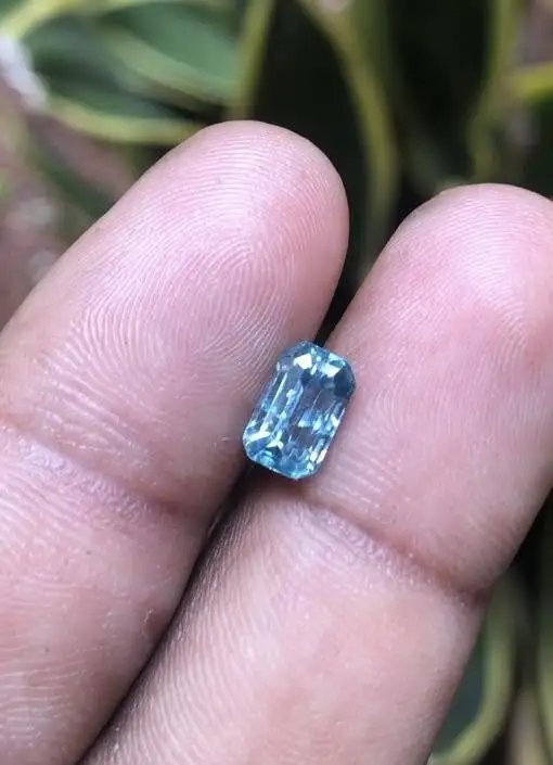 A person holding a small, faceted emerald-cut blue zircon gemstone between their fingers against a natural leafy background.